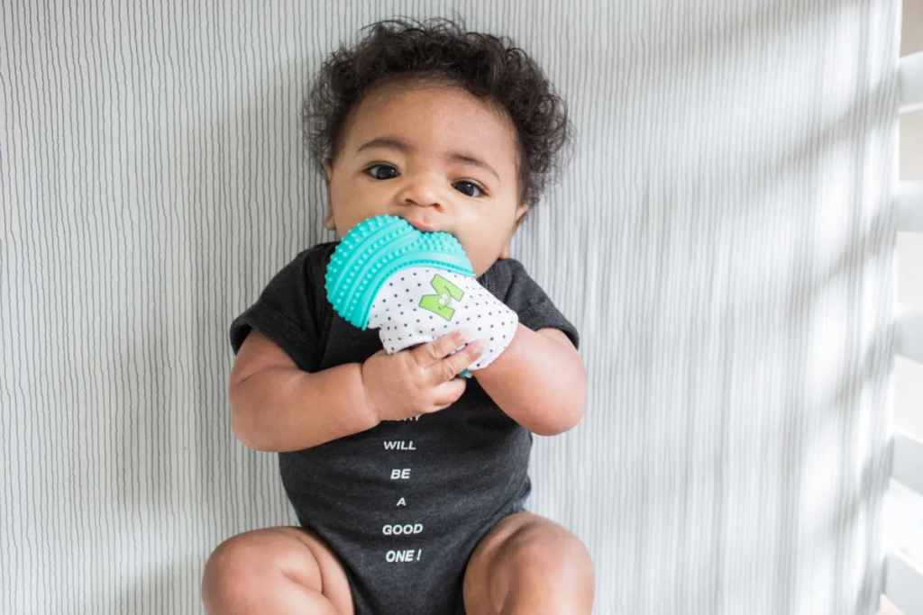 Baby wearing a 3-in-1 teething mitten, happily chewing on the soft, textured surface for teething relief, with a secure strap to keep it in place.